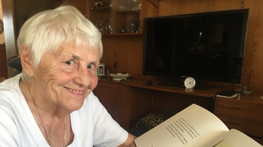 Läöchelnde, weißhaarige Dame sitzt im Wohnzimmer mit Buch über 2.Weltkrieg