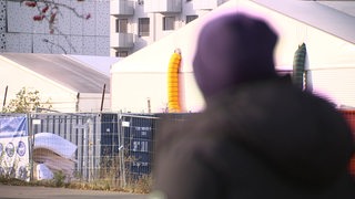 Im Hintergrund ist eine Flüchtlingsunterkunft scharf zu erkennen, im Vordergrund unscharf und von der Kamera abgewandt, eine Person mit Mütze.
