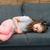 Kleines Mädchen liegt mit Bauchschmerzen auf einem grauen Sofa.