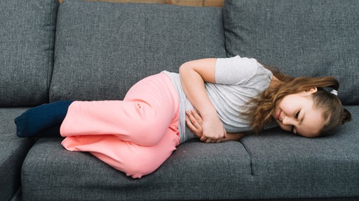 Kleines Mädchen liegt mit Bauchschmerzen auf einem grauen Sofa.