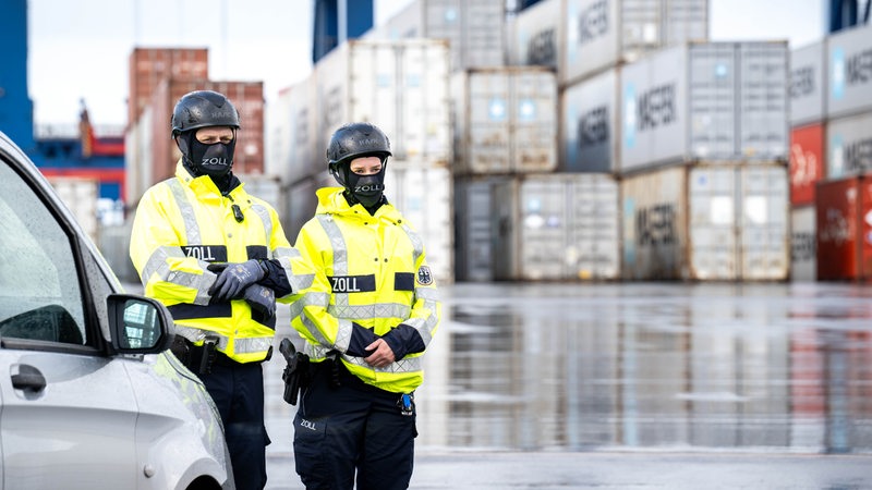 Zwei Mitarbeiter des Zolls stehen in gelben Jacken vor Containern in Bremerhaven