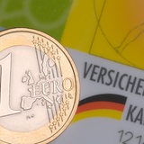 Eine Ein-Euro-Münze ist abgebildet. Im Hintergrund ist eine Krankenkassenkarte zu sehen.