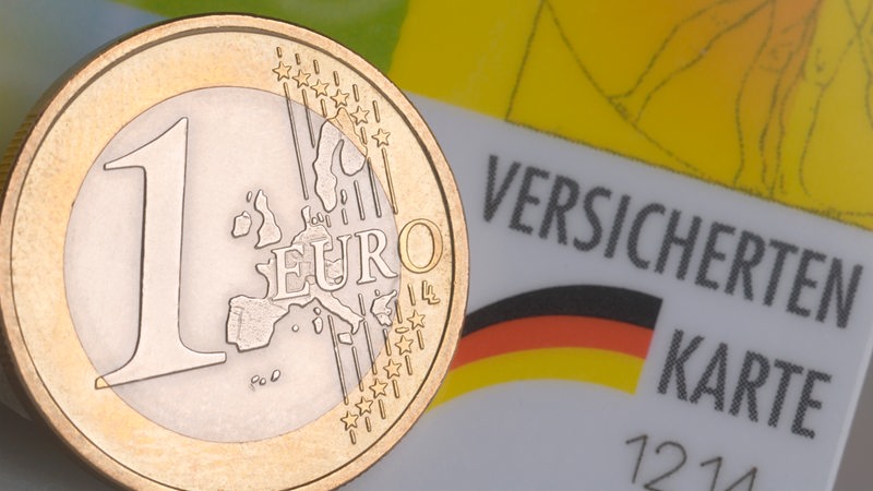 Eine Ein-Euro-Münze ist abgebildet. Im Hintergrund ist eine Krankenkassenkarte zu sehen.