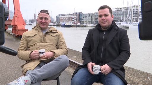 Zwei Männer sitzen vor einem Hafen und nehmen sich scheinbar mit einer Kamera auf.