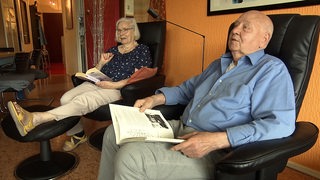 Eine Frau und ein Mann sitzen nebeneinander auf Sesseln in ihrer Wohnung.