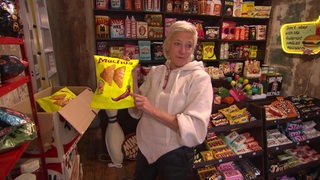Die Künstlerin Sylke Thoss steht in ihrem Kiosk und hält eine Chipstüte aus Pappmaché in den Händen.