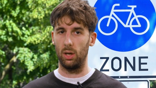 Ein Mann neben einem Schild "Fahrradzone" spricht in die Kamera.