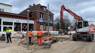 Auf einer Straße in Bremen-Sebaldsbrück stehen mehrere Bauarbeiter und ein Bagger vor einem Loch.