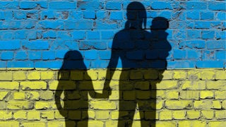 Auf eine blau-gelb gestrichene Wand sind die Schatten-Silhouetten einer Frau mit zwei Kindern gemalt.