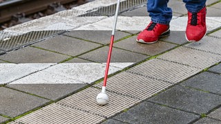 Ein Blindenstock mit einer Kugel ist auf einen Bahnsteig mit speziellen Bodenleitsystemen, auch Blindenleitsysteme genannt, zu sehen.