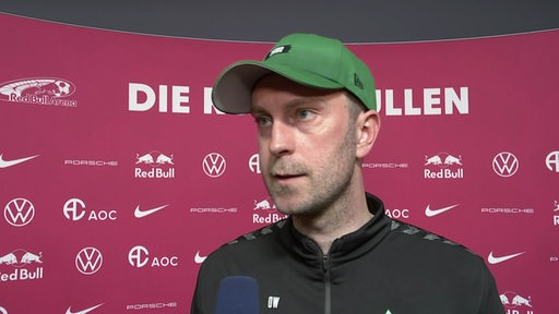Werder-Trainer Ole Werner mit grüner Kappe vor einer pinkfarbenen Werbewand beim Interview nach dem Spiel in Leipzig.