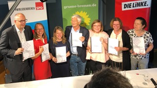 Sieben Personen stehen an einem Tisch und mit dem neuen Koalitionsvertrag zwischen SPD, Grünen und Linken.