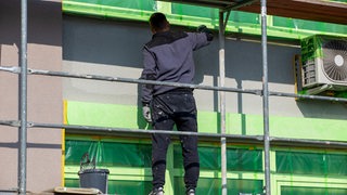 Ein Maler steht beim Streichen einer Fassade auf einem Gerüst.