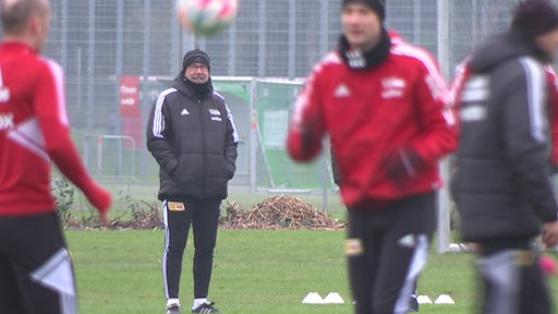 Union Berlin Trainer Urs Fischer am Spielfeldrand. Im Vordergrund wärmt sich seine Mannschaft beim Training auf.